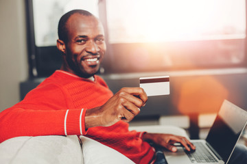 Portrait of pleased african man making purchase by internet while resting with notebook indoors. Focus hand holding credit card