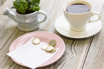 Pay restaurant bill. Bill, bank card, coins near cup of coffee on light wooden table top view