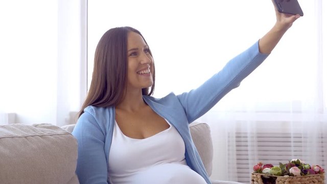 Positive pregnant woman making selfie on sofa in room
