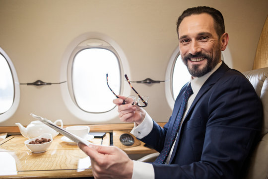 Portrait of contented successful man reading newspaper in plane, he is holding his glasses and looking at camera with smile