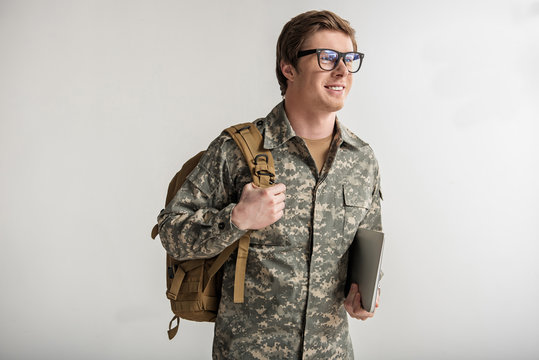 Happy male cadet holding laptop and smiling. He is wearing eyeglasses and having rucksack on shoulder. Isolated on background