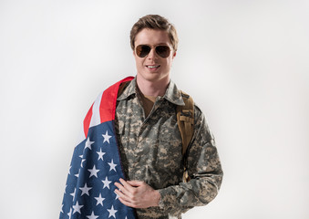 Waist up portrait of confident military young man looking at camera and smiling. American Flag is lying on his shoulder and he is having rucksack behind his back. Isolated on background