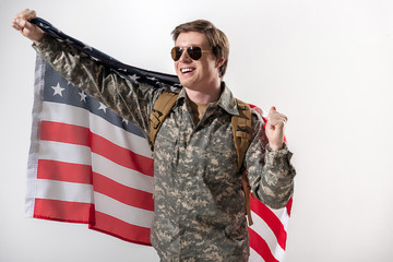 Joyful male soldier raising his hand while holding a usa flag behind his back. He is smiling and wearing eyeglasses. Isolated on background