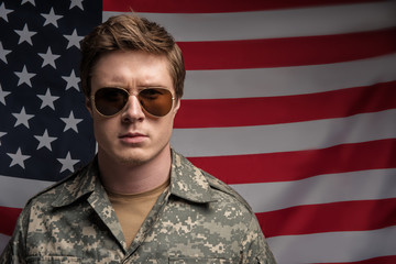 Portrait of earnest young officer looking at camera with serious look. He is wearing spectacles. Flag on background. Copy space