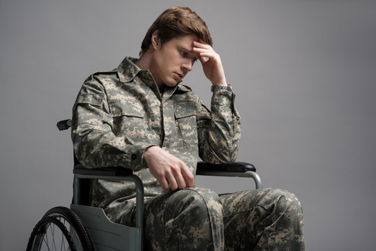 Hopeless young military man sitting in wheelchair. He is looking down with frustrated look and propping his head with his hand. Isolated on grey background