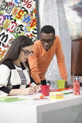 Two diverse work colleagues smiling and writing down notes while sitting together at a table in a modern office