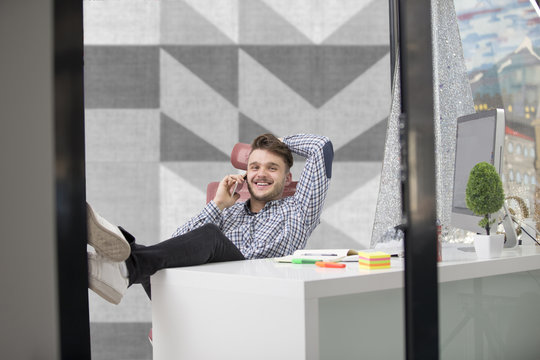 Handsome businessman in classic shirt is using a smartphone and smiling while sitting with legs on table in office