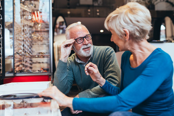 Happy senior couple choosing together eyeglasses frame in optical store.  - obrazy, fototapety, plakaty