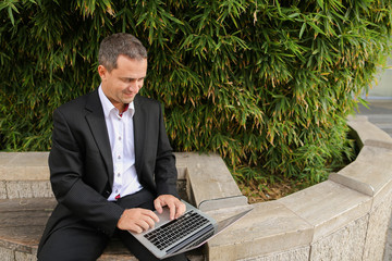 Businessman typing by laptop outside near green plant in   and looking at watch. Concept of ecology...