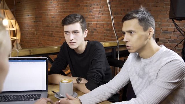 Group of designers are discussing their project in modern cafe. People are sitting at the wooden table with cup of coffee and brand-new laptop on. Young male professiona in white pulover is explaining