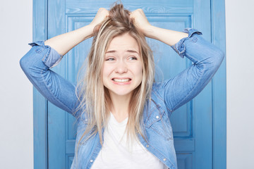 Isolated on blue door background young beautiful dressed casual Scandinavian woman is raging at herself, forgetting about important meeting, couldn’t turn back time, grimacing and clowning from anger.