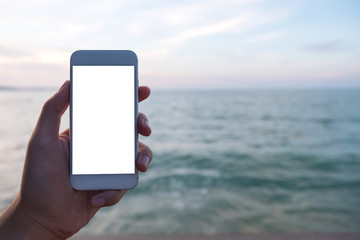 Mockup image of a hand holding and showing white mobile phone with blank desktop screen in front of the sea and sky background