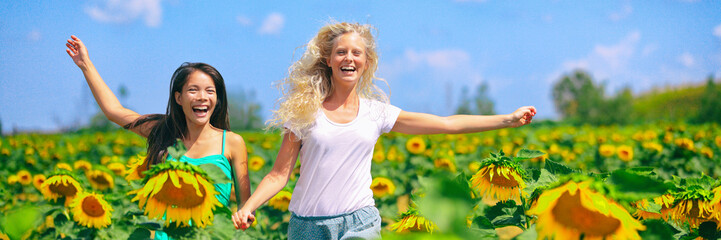 Two happy young women running through field of sunflowers on a sunny summer day laughing having fun with arms up in the fresh air. Beautiful young multiracial Asian and Caucasian girls banner.