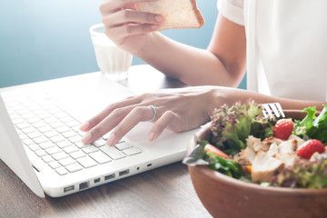 Obraz na płótnie Canvas Woman's hand using laptop computer and eating sliced bread with salad bowl in foreground
