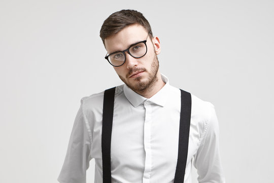 Serious handsome young unshaven male employee wearing trendy eyeglasses and white formal shirt with suspenders posing isolated against white studio wall background with copyspace for your information
