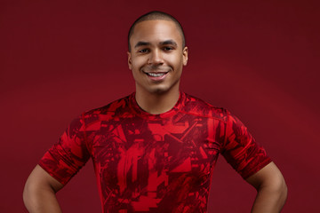 People, style, sports and fashion concept. Cropped shot of positive well set young African American male in his twenties posing in studio wearing trendy red t-shirt, looking and smiling at camera