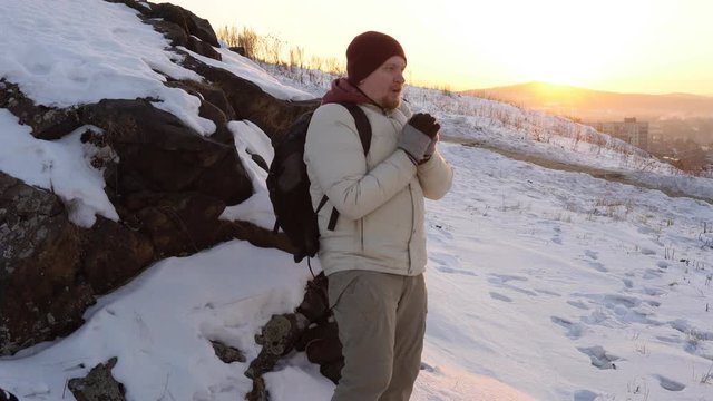 The tourist puts his hands in gloves to his mouth and exhales warm air, tries to warm up, in the snow-capped mountains near the city in winter at sunset.