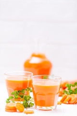 Carrot juice in beautiful glasses, cut orange vegetables and green parsley on white wooden background. Fresh orange drink. Close up photography. Selective focus. Vertical banner