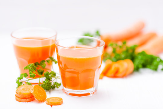 Carrot juice in beautiful glasses, cut orange vegetables and green parsley on white wooden background. Fresh orange drink. Close up photography. Selective focus. Horizontal banner