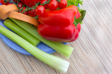 big celery,tape measure for diet, fresh green lettuce leaves next to red pepper and red cherry tomatoes lying on a blue plate, wooden table background