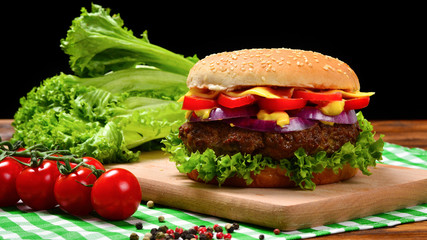 Delicious tasty burger with beef, tomatoes, salad and sauce on wooden table and black background.  Space for text or design.