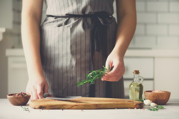 Woman hands cutting fresh green rosemary on wood chopping board in white kitchen, interior. Copy space. Homemade food conceplt, healthy recipe. Take me to work