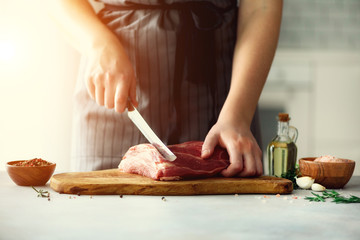 Obraz premium Wooman hands cutting beef meat on wooden chopping board, rosemary, oil, salt, pepper. Girk cooking pork meat on white kitchen backgound. Copy space