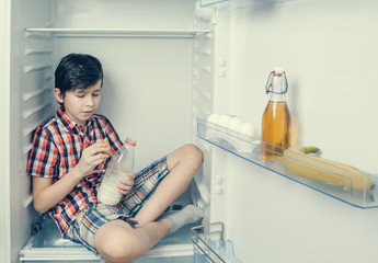 A boy in a shirt and shorts eating a croissant and drink milk inside a fridge  with food and product.