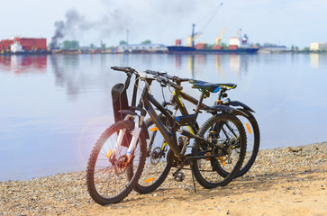 Fototapeta premium The couple of bicycles are next to a river in the urbanistic landscape. The parked bikes are a riverbank at the background of urban area.