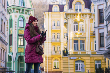 Fototapeta premium Pretty Smiling Brunette Girl Wearing Purple Winter Coat, Hat and Scarf, Walking by European Street at Winter