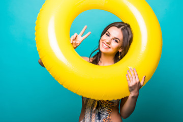 Close up portrait of a beautiful woman in bikini look through big yellow inflatable ring and show victory sign to the camera on green background