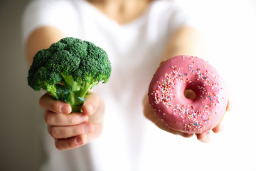 Young woman in white T-shirt choosing between broccoli or junk food, donut. Healthy clean detox...