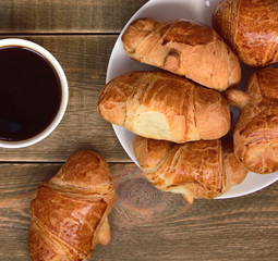  coffee mood. close up of a cup of black coffee and a plate with freshly baked croissants with golden crust and one croissant is next to a cup on a brown wooden table