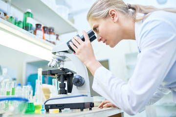 Side view portrait of young female scientist looking in microscope while working on medical...