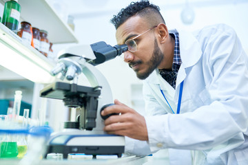 Side view portrait of young Middle-Eastern scientist looking in microscope while working on medical...