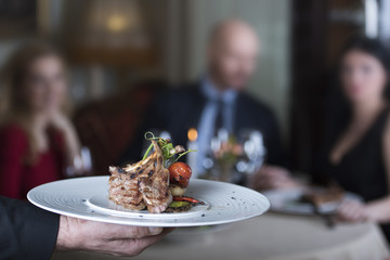 Delicious grilled lamb ribs, served with grill tomato and decorated with radish, baby spinach and green leafs, placed on white plate, presented to a group of persons - unrecognizable faces, restaurant