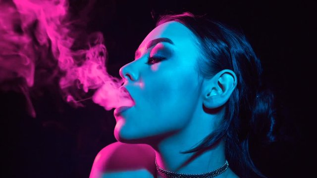 Portrait of glamour seductive gorgeous brunette woman smoking electronic cigarette in neon color light in studio