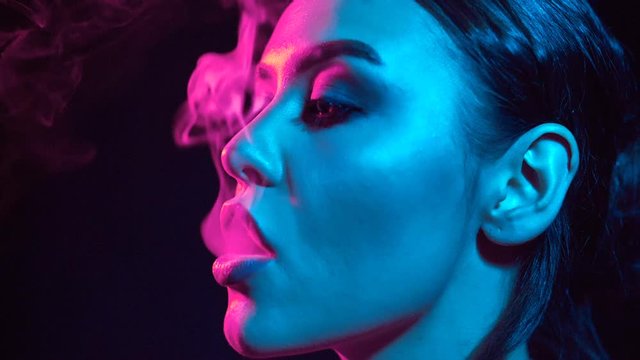 Portrait of glamour seductive gorgeous brunette woman smoking electronic cigarette in neon color light in studio