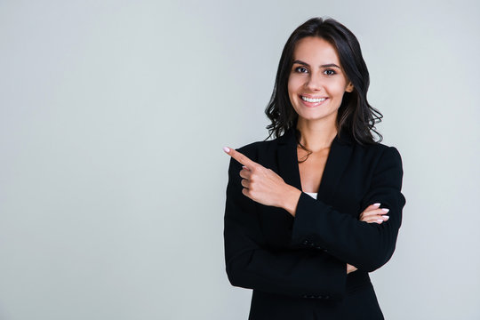 You need to look here! Beautiful young businesswoman pointing away and looking at camera with smile while standing against white background