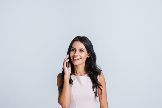 Such A Nice Talk. Beautiful Young Woman Talking On Her Smartphone And Looking At Away With Smile While Standing Against White Background