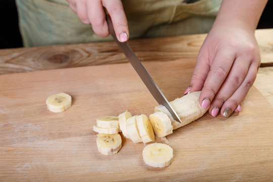 On a wooden table on a cutting board, a woman steers a banana knife to pieces. Middle plan, front view.