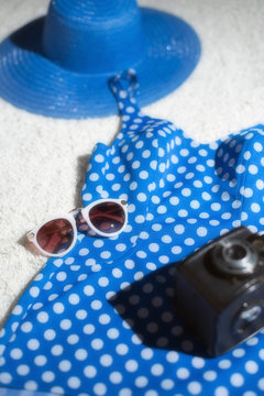 Blue vintage hat with swimsuit, camera and sunglasses displayed on white carpet floor.