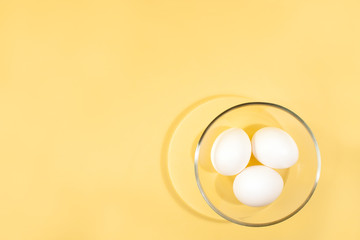 Three white eggs are lying in a round transparent glass cup in the right bottom of a yellow background.