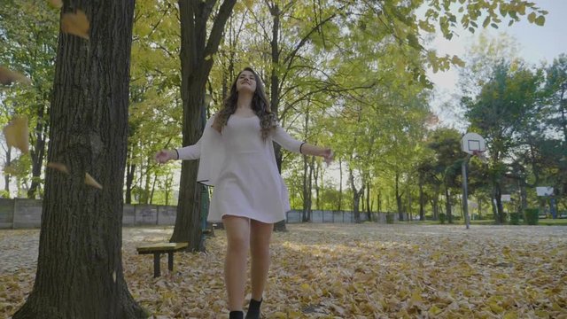 Happy young girl in white clothes walking in the park admiring falling autumn yellow leaves and enjoying freedom in slow motion
