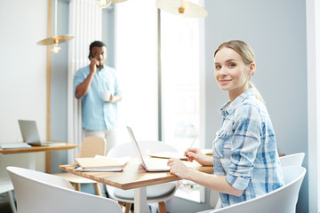Obraz premium Happy young businesswoman sitting by table in office and preparing for report with man calling on background