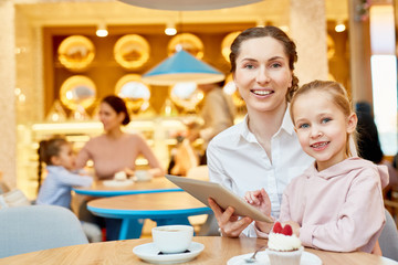 Young smiling female and her cute daughter sitting by table in cafe, having dessert with tea and watching online movie
