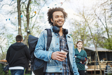 Outdoor portrait of handsome unshaved dark-skinned guy standing in park with coffee, wearing headphones over neck and looking aside. Man tries to find his girlfriend in crowd.