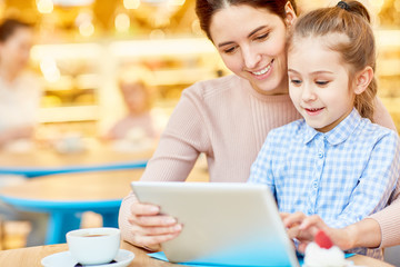Happy young woman and her daughter sitting in cafe with tablet and having fun while searching in the net