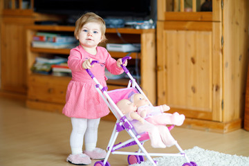 Cute adorable baby girl making first steps with doll carriage.