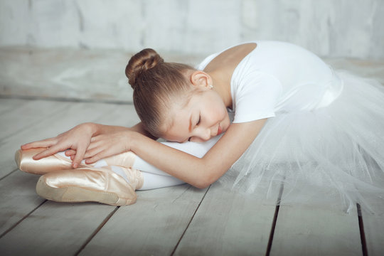 A little adorable young ballerina in white dress and point shoes lies lika a swan on woody floor and gray studio background posing on camera